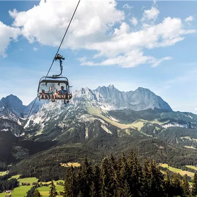 A cable car glides over green meadows and high mountains. The sky is clear and blue with some clouds.