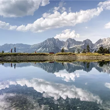 An idyllic mountain landscape with clear, reflecting waters. In the background, impressive mountains and a stunning cloud formation can be seen.