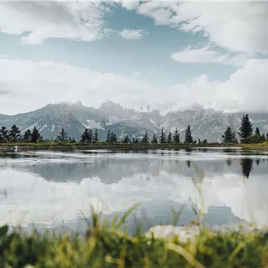 Ein ruhiger See im Vordergrund spiegelt die umliegenden Berge und den bewölkten Himmel wider. Grüne Wiesen und Bäume rahmen die Szenerie ein.
