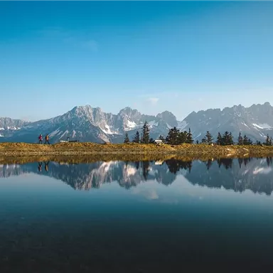Eine malerische Berglandschaft mit schneebedeckten Gipfeln und einem ruhigen See im Vordergrund. Zwei Wanderer genießen die schöne Aussicht.