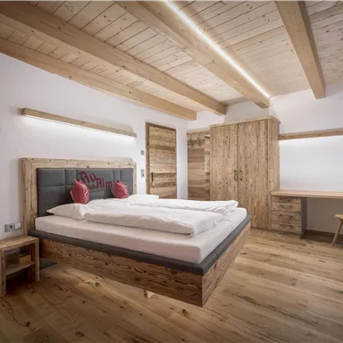 A modern bedroom with wood decor and a large bed. The room is bright and friendly with a desk and a wardrobe.