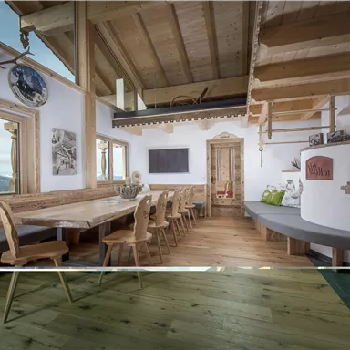 A cozy room with wooden interior and a large dining table. In the background, mountain landscapes can be seen through the windows.