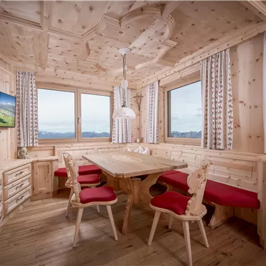 A cozy dining area made of light wood with a large table and red chairs. The windows offer a beautiful view of the mountains.