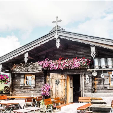 A cozy wooden house with colorful flowers and Bavarian decoration. In front of the house, there are tables and chairs in a beautiful outdoor area.