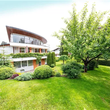 Ein modernes Haus mit einer einzigartigen Dachform, umgeben von grünem Rasen und Bäumen. Die gepflegte Gartenlandschaft sorgt für eine einladende Atmosphäre.