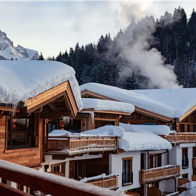 An idyllic mountain cabin in the snow with wooden clad facades. In the background, there are snow-covered mountains and rising smoke.