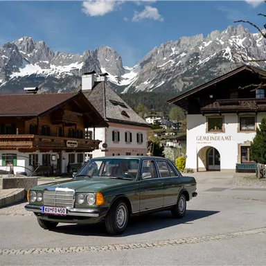 Ein elegantes Auto fährt durch ein malerisches Dorf mit traditionellen Gebäuden. Im Hintergrund sind majestätische Berge und eine klare Himmelsaufnahme zu sehen.