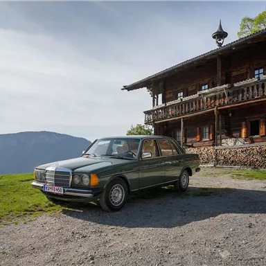 Ein klassisches Auto steht vor einem traditionellen Holzhaus in einer landschaftlichen Umgebung. Im Hintergrund sind Berge und ein klarer Himmel zu sehen.