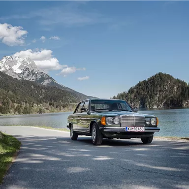 Ein klassisches Auto parkt an einer malerischen Straße entlang eines klaren Sees. Im Hintergrund sind Berge und ein blauer Himmel zu sehen.