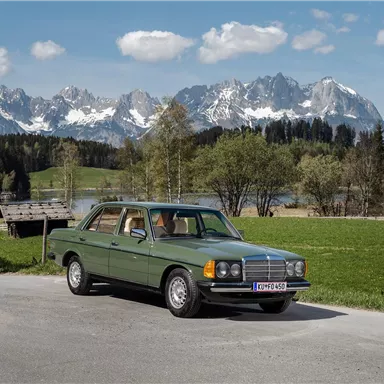 Ein klassisches Auto steht auf einer Straße vor einer malerischen Berglandschaft. Im Hintergrund sind schneebedeckte Berge und grüne Wiesen zu sehen.