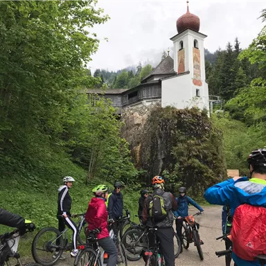 Eine Gruppe von Radfahrern steht auf einem Weg, umgeben von üppigem Grün. Im Hintergrund ist eine kleine Kirche mit einer markanten Kuppel zu sehen.