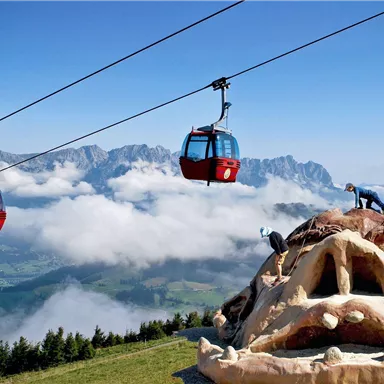 A cable car glides over an impressive mountain landscape. Two people are climbing on a large artwork in the shape of a face.