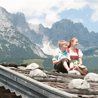 Zwei Kinder sitzen auf einem Dach und genießen die Aussicht. Im Hintergrund sind majestätische Berge und ein blauer Himmel zu sehen.
