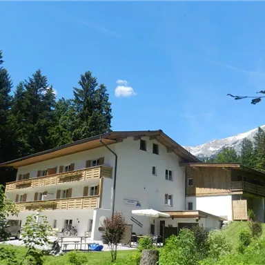 Ein gemütliches Gebäude umgeben von grünen Bäumen und einem klaren Himmel. Im Hintergrund sind Berge sichtbar, was eine ruhige, alpine Atmosphäre schafft.