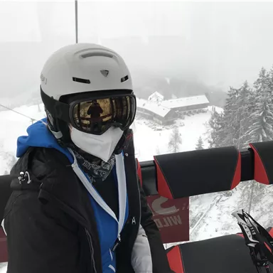 Eine Person in Skiausrüstung sitzt in einer Gondel auf dem Weg zum Skigebiet. Im Hintergrund sind schneebedeckte Bäume und ein Haus zu sehen.