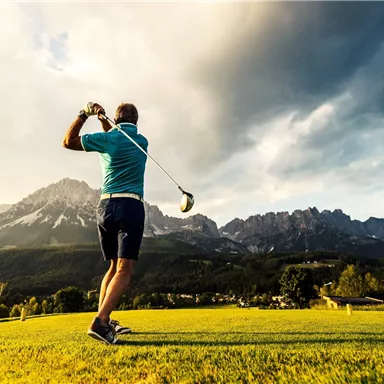 A man is playing golf on a course with impressive mountains in the background. The sky is overcast, creating a dramatic atmosphere.