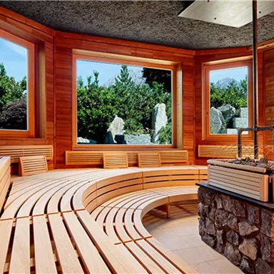 Eine helle Sauna mit Holzbänken und großen Fenstern. Draußen sieht man üppige Grünanlagen und Felsen.