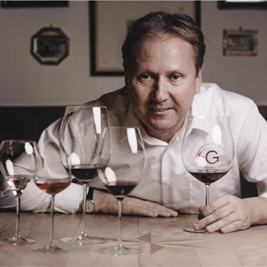 A man is sitting at a table with several wine glasses in front of him. He is holding a glass of red wine and looking straight into the camera.