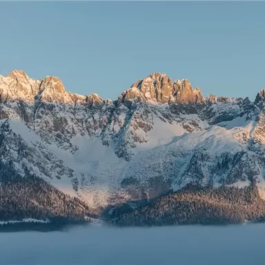 An impressive mountain landscape with snow-capped peaks and a gentle mist in the valley. The sky is clear and glows in warm morning colors.