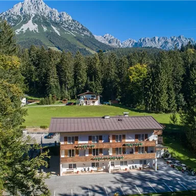 Ein malerisches Hotel umgeben von Bergen und Wäldern. Vor dem Hotel befindet sich ein Parkplatz und eine grüne Wiese.