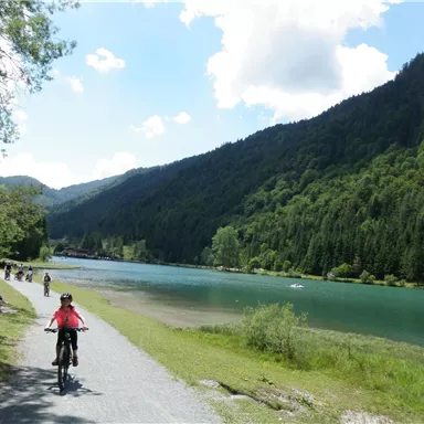 Ein Radfahrer fährt auf einem Weg entlang eines klaren, blauen Sees. Im Hintergrund erheben sich grüne Berge unter einem strahlend blauen Himmel.