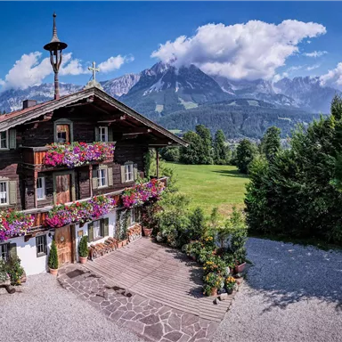 A charming wooden house with colorful flowers on the windowsills stands in front of an impressive mountain backdrop. The sky is clear, and the nature around the house is green and inviting.