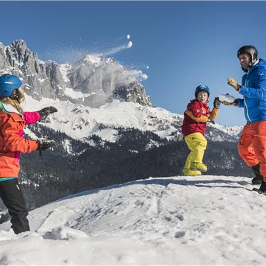 Eine Gruppe von drei Menschen mit Skikleidung hat Spaß im Schnee. Im Hintergrund sind beeindruckende Berge unter einem klaren blauen Himmel zu sehen.