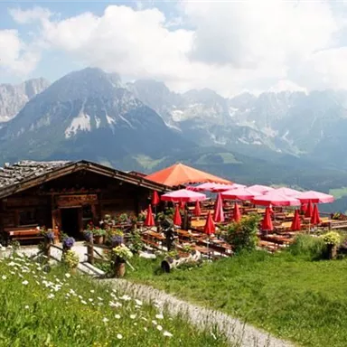 Eine malerische Berghütte mit Terrasse und roten Sonnenschirmen. Im Hintergrund sind beeindruckende Berge und ein blauer Himmel zu sehen.
