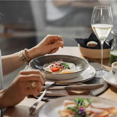 Ein elegant gedeckter Tisch mit verschiedenen Gerichten und einem Glas Wein. Eine Hand hält Besteck, während eine Tasse Kaffee und frische Blumen im Hintergrund zu sehen sind.