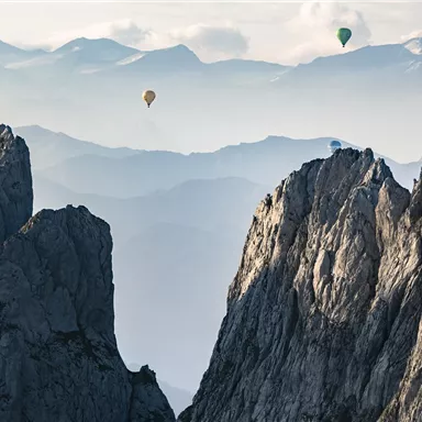 Heutzutage sieht man Luftballons über einer beeindruckenden Berglandschaft. Die schroffen Gipfel heben sich vor einem sanften, nebligen Hintergrund.
