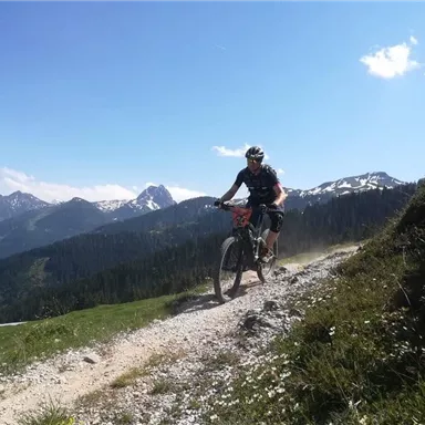 Ein Radfahrer fährt auf einem Schotterweg durch die Berge. Der Himmel ist klar und die Landschaft ist grün und bergig.
