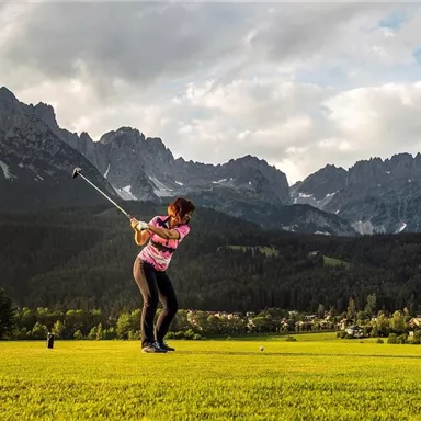 Eine Golferin schlägt in einer malerischen Landschaft mit Bergen im Hintergrund. Die grüne Wiese und der bewölkte Himmel schaffen eine eindrucksvolle Kulisse.