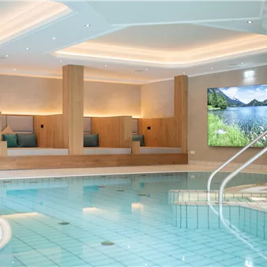 A relaxing wellness area with a swimming pool and cozy loungers. On the wall, there is a screen displaying a landscape image.