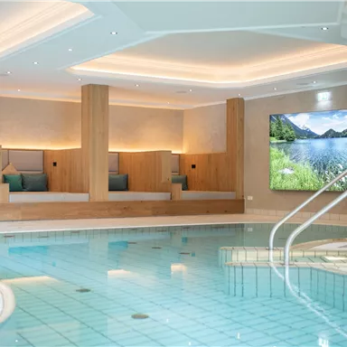 An elegant indoor pool with smooth water and modern loungers. In front of a wall, there is a large screen displaying a picturesque landscape.