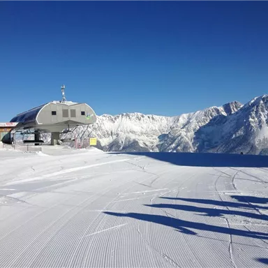 Eine schneebedeckte Berglandschaft mit einem Lifthaus im Vordergrund. Im Hintergrund sind majestätische Berge unter einem klaren blauen Himmel zu sehen.