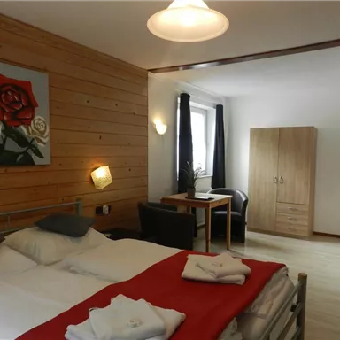 Ein gemütliches Zimmer mit einem Doppelbett, einem Tisch und Stühlen. Die Wände sind teilweise holzverkleidet und ein Kunstwerk mit Rosen schmückt den Raum.