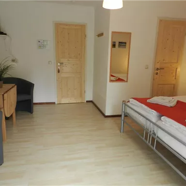 Ein einfaches Zimmer mit einem Doppelbett, einem Tisch und einem Stuhl. Die Wände sind hell und es gibt viel Tageslicht.