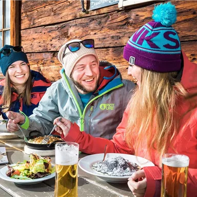 Eine Gruppe von drei Freunden sitzt entspannt an einem Tisch in einer Berghütte. Sie genießen Essen und Getränke in einer gemütlichen Atmosphäre.