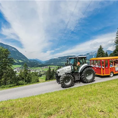 A tractor is pulling a colorful wagon with passengers on a winding road. In the background, green hills and a clear sky can be seen.