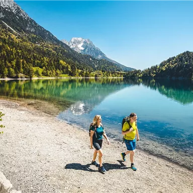 Zwei Wanderer spazieren an einem klaren, blauen See. Im Hintergrund sind grüne Wälder und Berge zu sehen.
