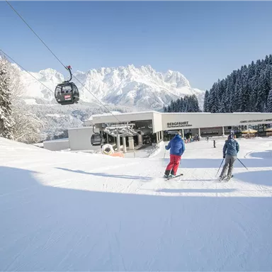 Eine winterliche Landschaft mit schneebedeckten Bergen im Hintergrund. Zwei Skifahrer genießen die Piste in der Nähe einer Gondelbahn.