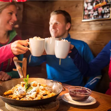 Three friends toast with mugs and enjoy a meal in a cozy setting. On the table, there's a plate with delicious treats.
