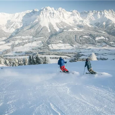 Two skiers are skiing on a snowy slope with impressive mountain panoramas in the background. The sky is clear and the landscape is beautifully wintry.