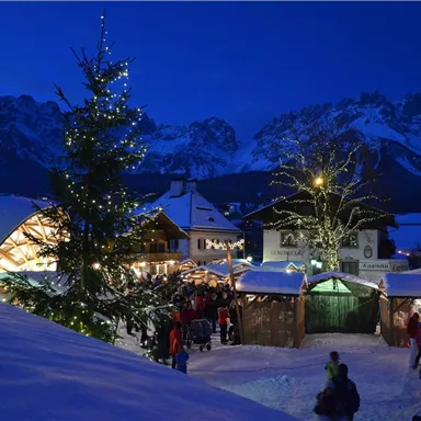 Ein festlicher Weihnachtsmarkt in einer verschneiten Berglandschaft. Die Stände sind beleuchtet und es gibt einen geschmückten Weihnachtsbaum.