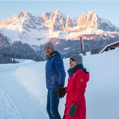 A couple is walking hand in hand in the snow. In the background, majestic mountains and a clear sky can be seen.