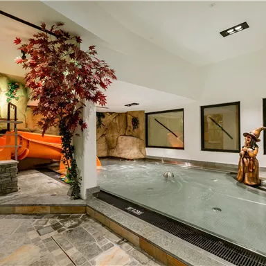 A wellness area with a small pool and a waterslide. In the background stands a statue, surrounded by plants.