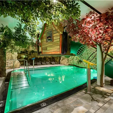 A stylish indoor pool surrounded by green plants and trees. A small wooden cabin and a slide create a relaxed atmosphere.