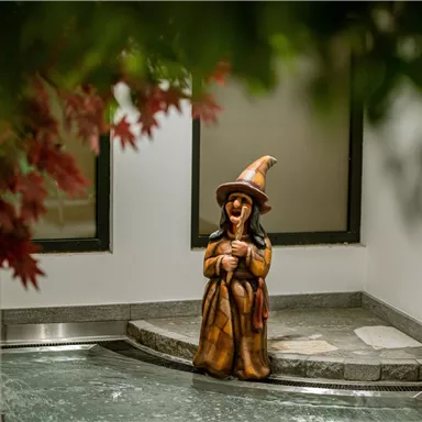 A wooden figure of a witch stands in a hallway. She is wearing a hat and smiling.