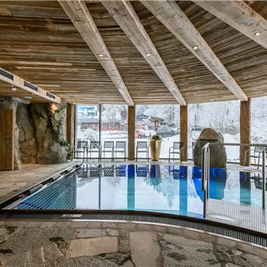 An elegant indoor pool with a wooden ceiling and stone walls. A snowy landscape can be seen outside the window.