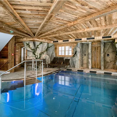 A modern indoor pool in a rustic wooden room. The walls are partly designed with rocks and large windows provide plenty of light.
